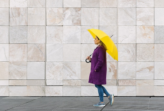 Blonde woman wearing a purple coat walking with a yellow umbrella on a stone architecture background.