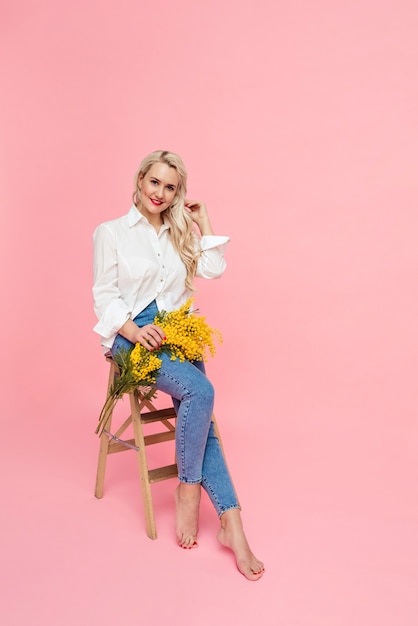 A blonde woman in a white shirt and jeans holds yellow flowers on a pink background