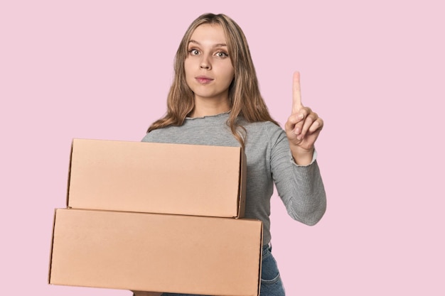 Blonde woman with cardboard boxes moving on studio backdrop showing number one with finger