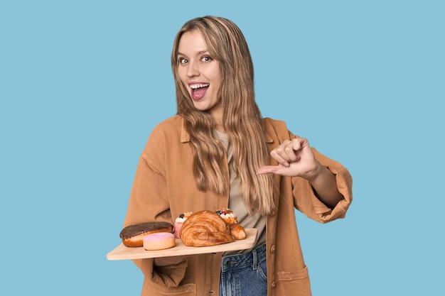 Blonde woman with pastry tray in studio backdrop points with thumb finger away laughing and carefree