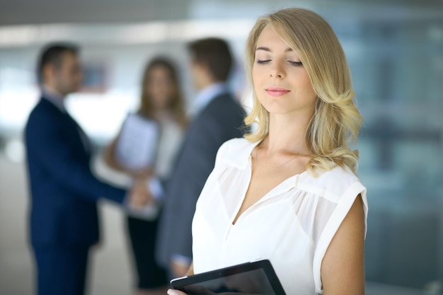 Blonde woman with touchpad computer looking at camera and smiling while business people shaking hands over background