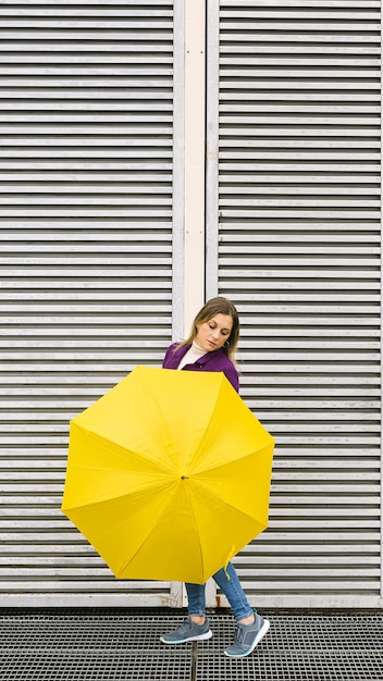 Blonde woman with a yellow umbrella posing in front of a white wall with a geometric structure.