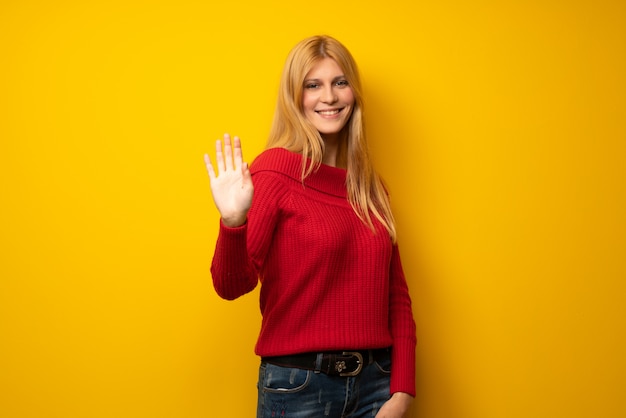 Blonde woman over yellow wall saluting with hand with happy expression