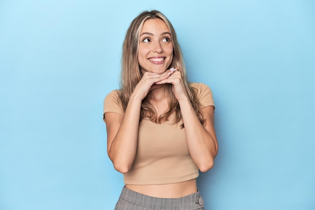 Blonde young caucasian woman in blue studio keeps hands under chin is looking happily aside