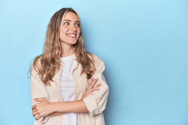 Blonde young caucasian woman in blue studio smiling confident with crossed arms