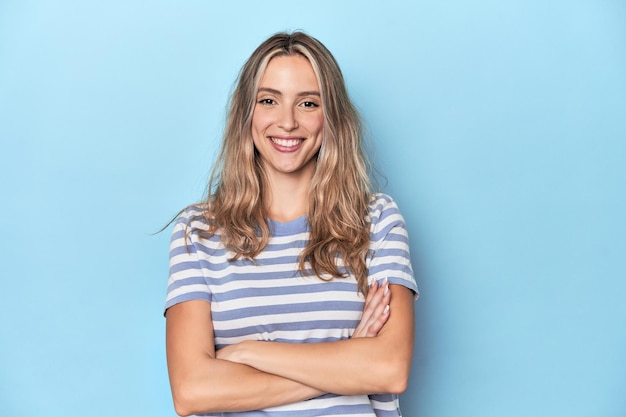 Blonde young caucasian woman in blue studio who feels confident crossing arms with determination