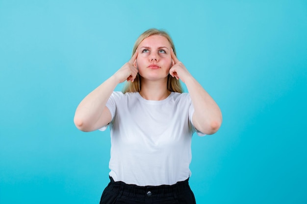 Blonde young girl is looking up by holding forefinger on temples on blue background