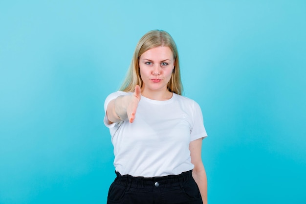 Blonde young girl is showing hi gesture by extending hand to camera on blue background