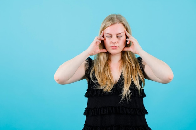 Blonde young girl is thinking by closing eyes and holding forefingers on temples on blue background