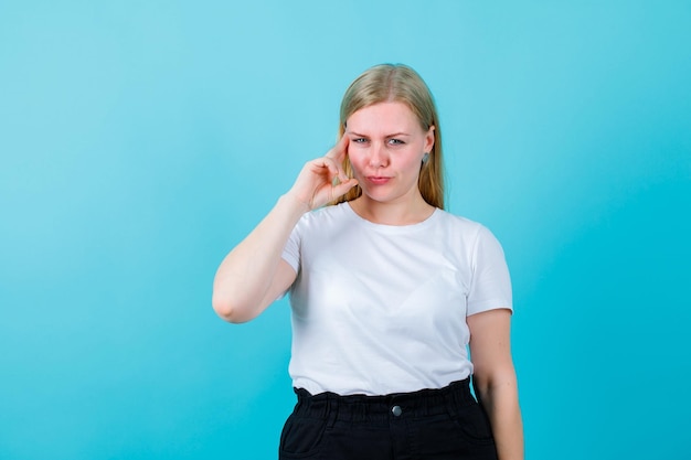 Blonde young girl is thinking by holding fingers on temple on blue background