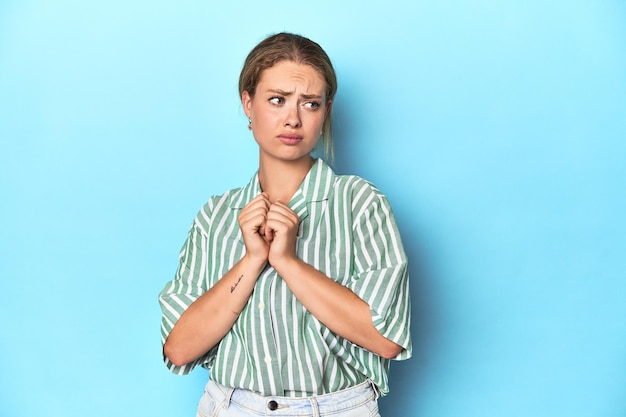 Photo blonde young woman in green striped shirt on blue background scared and afraid