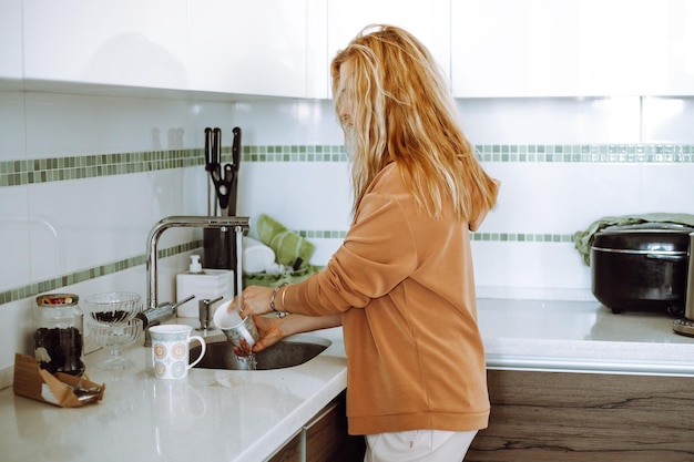 Blonde young woman in home clothes wash mugs in sink under running water after eating and tidy up mo