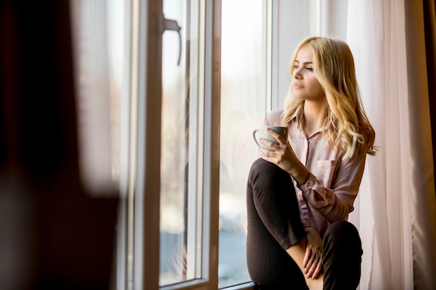 Blonde young woman relaxing and  drinking coffee by window