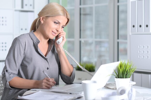 Blonde young woman working with laptop while talking on phone