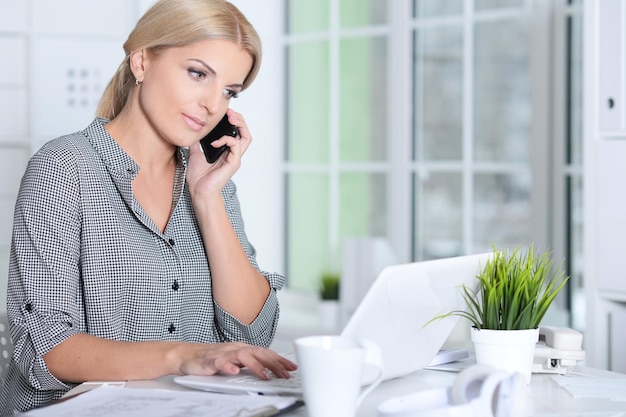 Blonde young woman working with laptop while talking on phone