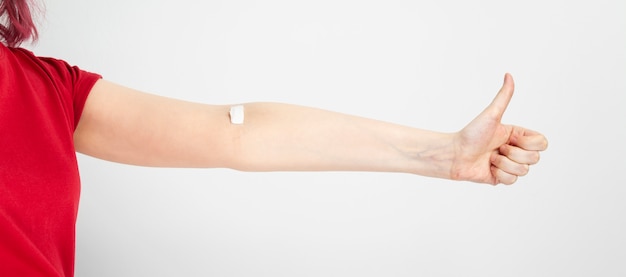 Blood donorship. Young girl in red T-shirt shows thumb up, second hand taped with patch after giving blood on grey background