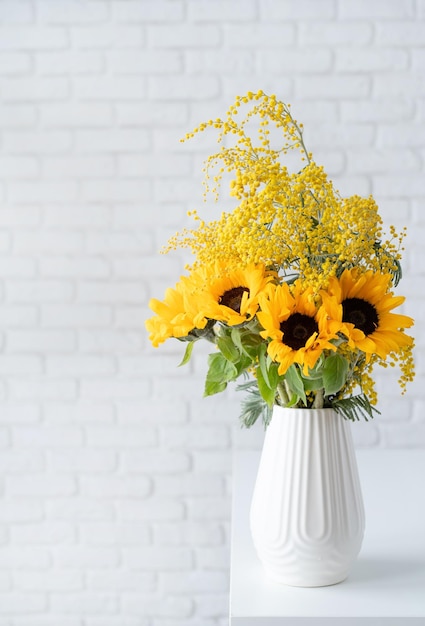 Blossom of Mimosa and sunflowers in white ceramic vase on white brick wall background with space for text