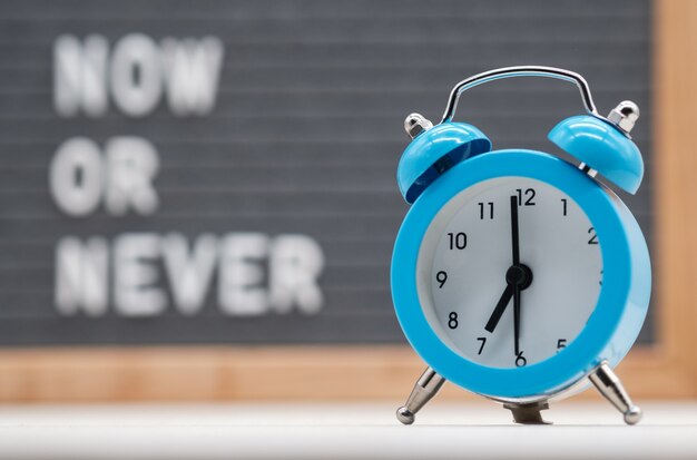 Photo blue analog alarm clock on english text background now or never . the concept of immediate action