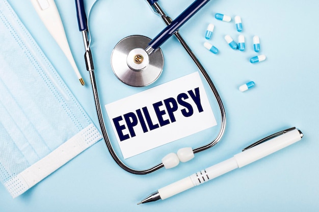 On a blue background a stethoscope a medical mask pills a pen and a white card with the text EPILEPSY View from above