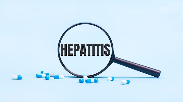 On a blue background white and blue capsules with pills and a black magnifying glass with the text HEPATITIS Medical concept