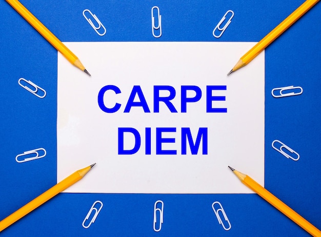 On a blue background white paper clips yellow pencils and a white sheet of paper with the text CARPE DIEM