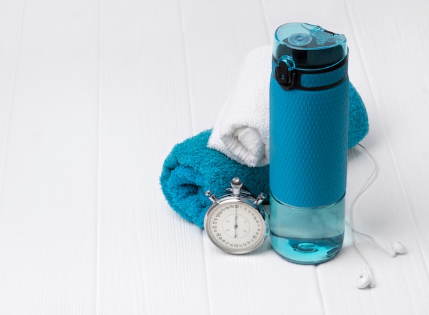 Blue bottle of water, towels, headphones and stopwatch. Sports equipment on white wooden table