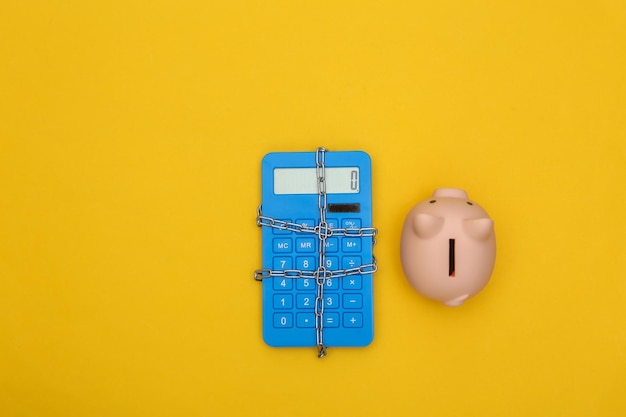 Blue calculator wrapped in steel chain and piggy bank on yellow background. Top view