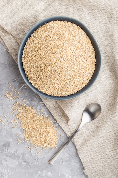 Blue ceramic bowl with raw white quinoa seeds on a gray concrete  background. Top view, close up.