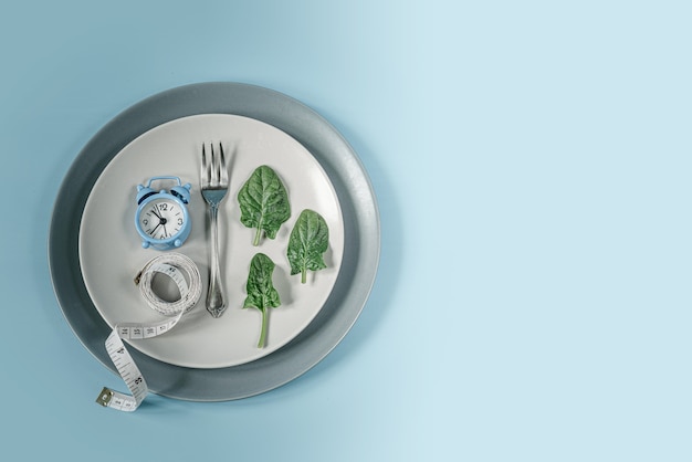 Blue clock, fork, spinach leaves and measure tape on gray plate, diet and intermittent fasting concept