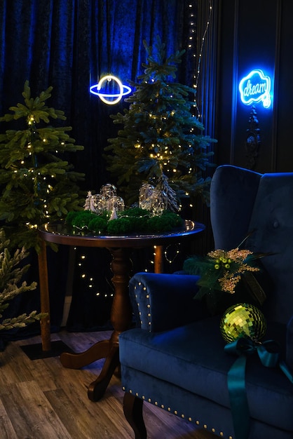 Blue and green Christmas interior. A fairy interior with a green wall, blue armchair, round table on which there are glowing bulbs decorated with moss and Christmas trees with lights and neon signs