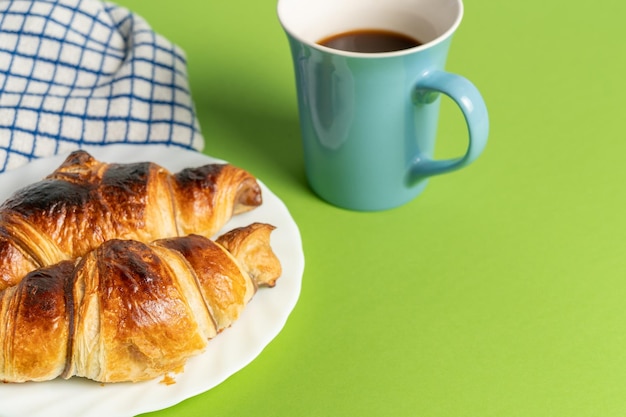 A blue mug of coffee sits next to a croissant on a green table.