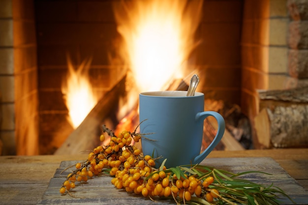 Blue mug of tea and a branch of yellow sea buckthorn near a cozy fireplace