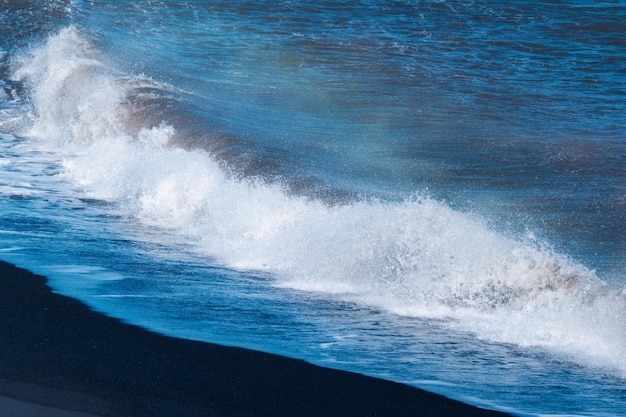 Blue ocean wave with splashing hitting on black sand beach in summer