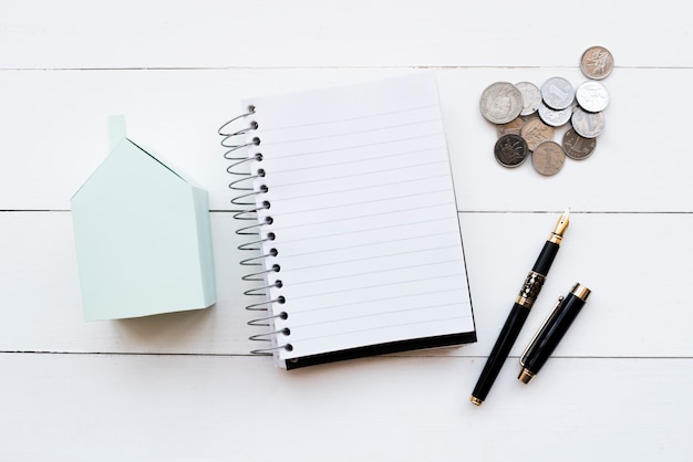 Blue paper house model; spiral diary; coins and black fountain pen with an open lid on white table