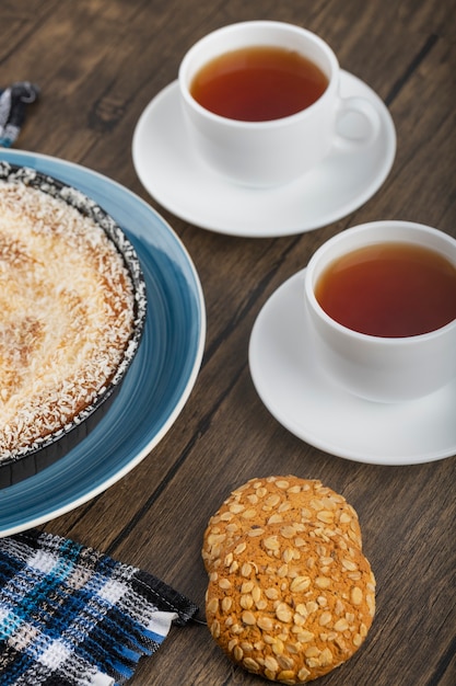 Blue plate of sweet pie with coconut sprinkles, cup of tea and biscuits on wooden surface