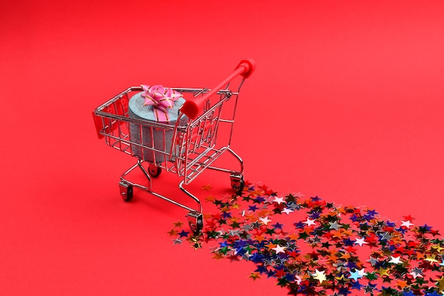 Blue present box with pink bow in a shopping cart and confetti on a red background