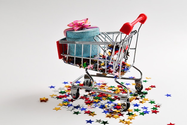 Blue present box with pink bow in a shopping cart and confetti on a white background.