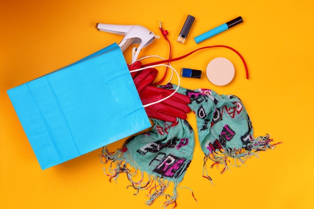 Blue shopping bag with stylish shoes, scarf, red belt and gloves and cosmetics on yellow background