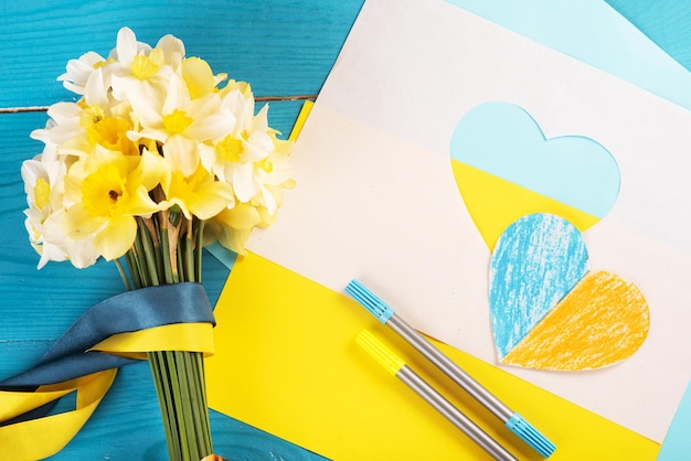 Blue and yellow color paper texture background heart cut out of paper and painted in blue and yellow colors and yellow spring flowers on wooding back Creativity as support of Ukraine Trend colors