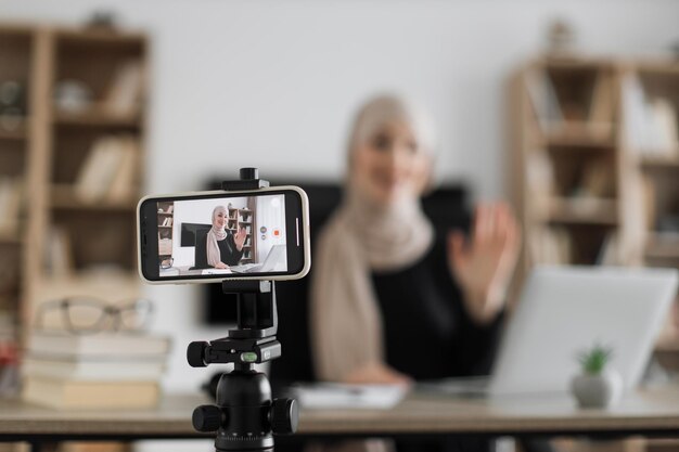 Blur background of pretty muslim woman with headscarf sitting at desk and filming video blog