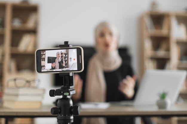 Blur background of pretty muslim woman with headscarf sitting at desk and filming video blog