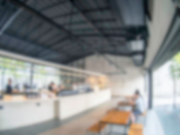 Blurred modern coffee shop or cafe or restaurant interior background Blurry white and bright coffee bar decoration with wooden table and chair near large glass window with sunlight from outside