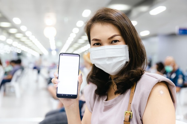 Blurred out of focus woman wearing hygiene protective face mask and show blank white screen smartphone while sitting in the indoor hall with bokeh lights and defocused people in background.