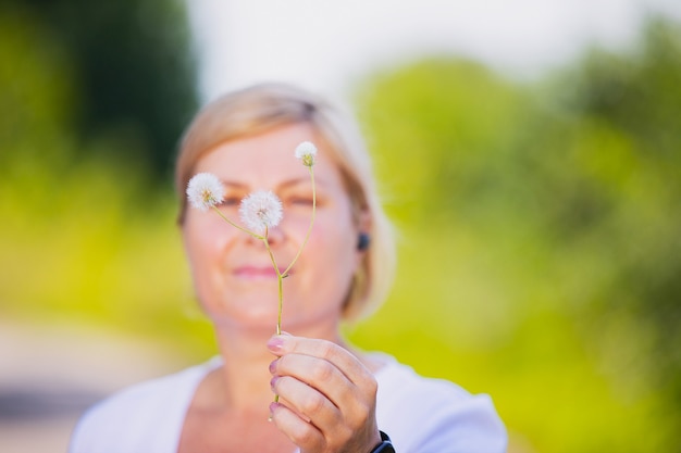 Blurred photo of a blonde woman outdoors holding the dandelion flower in her hand and wearing smart ...