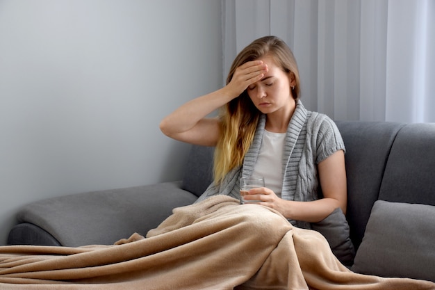 Blurred sick Caucasian woman with closed eyes suffer from fever on sofa is holding glass of water while touching her forehead with hand, feeling temperature. Illness, pain, disease, headache concept