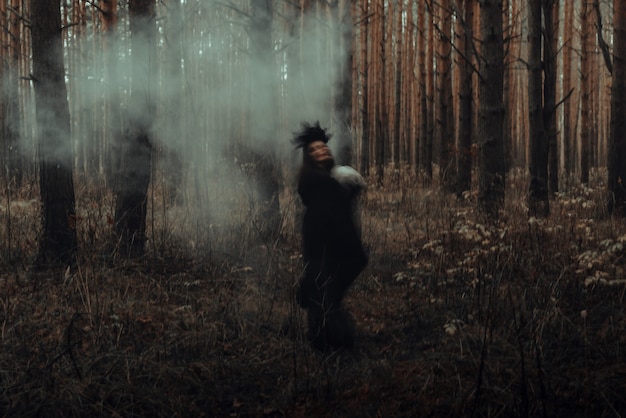 Blurred silhouette of a terrible witch with a skull in her hands performing a mystical occult satanic ritual in a dark gloomy forest