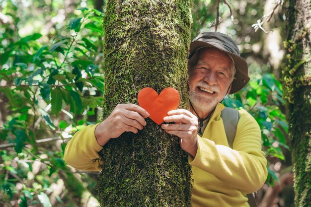 Blurred smiling senior man in the woods holding a paper heart on a moss covered tree trunk earth day