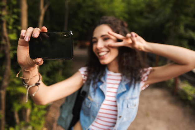 Blurry brunette teenage woman with backpack, showing peace sign and taking selfie photo on smartphone while walking in green park