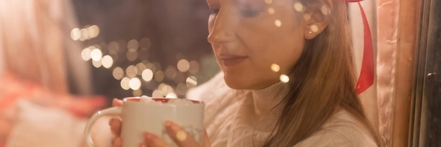 Blurry portrait candid woman face through window glass holding in hands and enjoyment cup of hot chocolate drink and relaxing on happy new year and merry christmas eve glowing lights garland banner