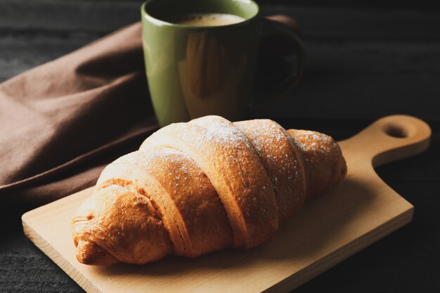 Board with croissant and cup of coffee on wooden background, close up